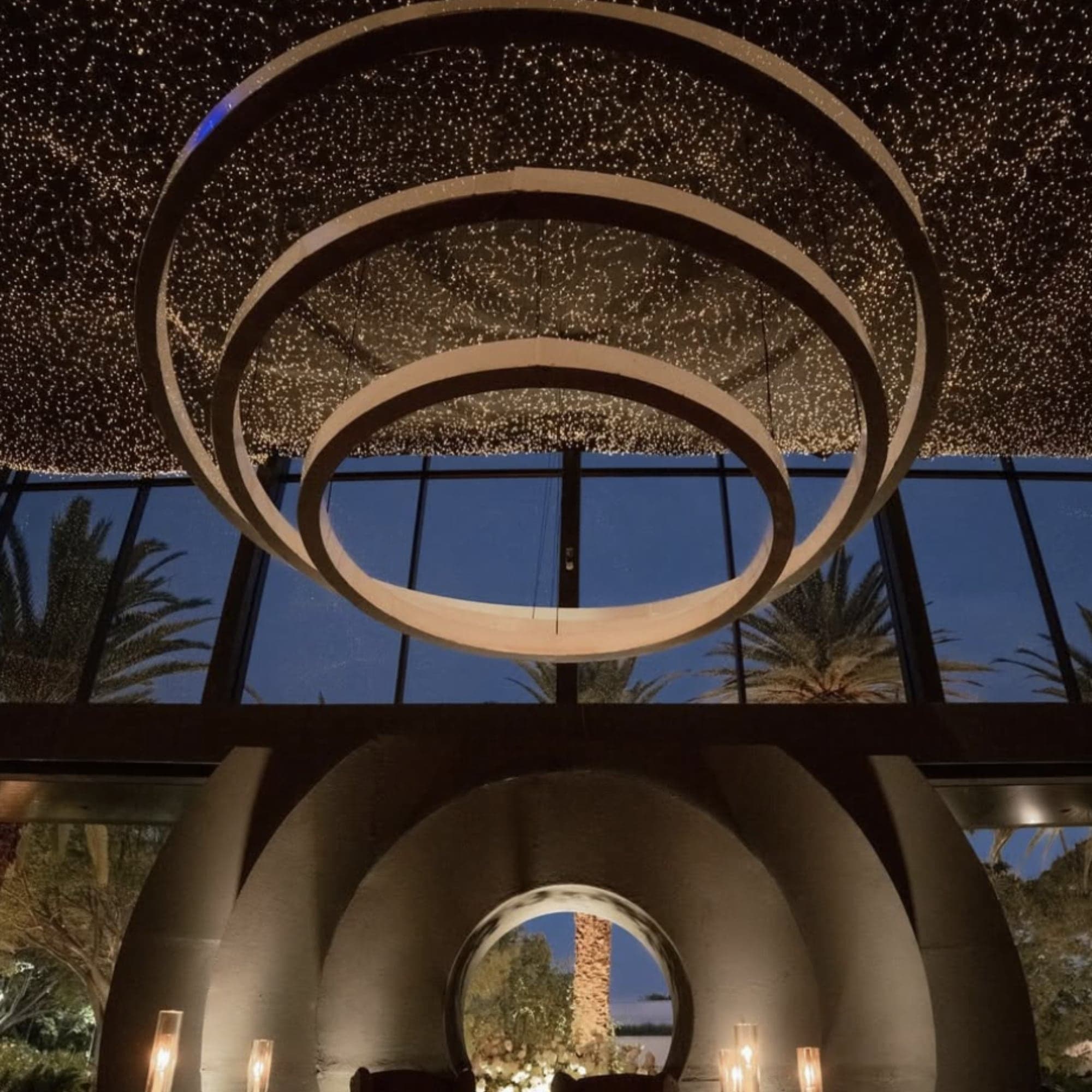 Lounge nocturno con candelabro de anillos concéntricos bajo un techo de luces y palmeras al fondo