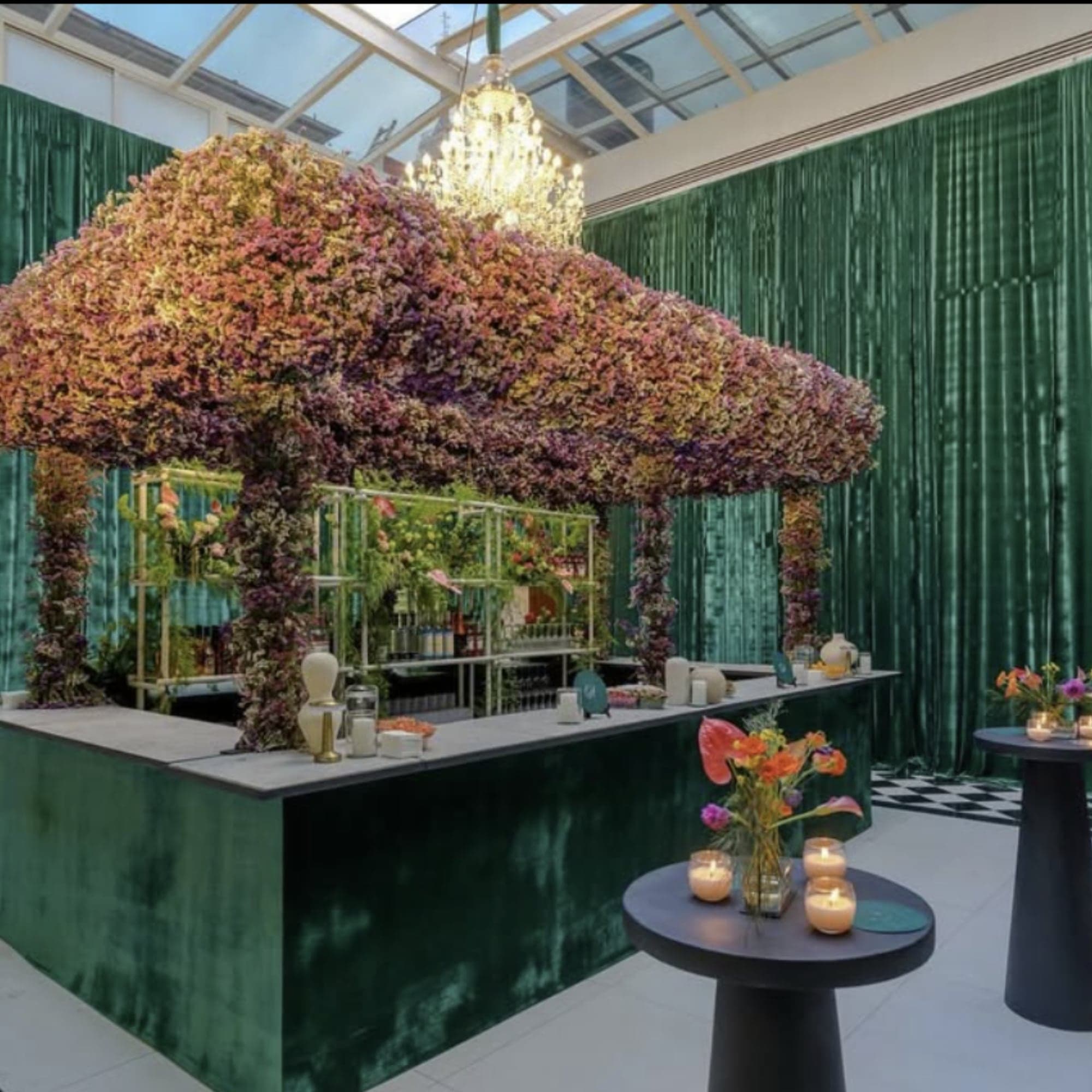 Barra de coctel bajo un dosel de flores, con cortinas de terciopelo verde y mesas altas con velas