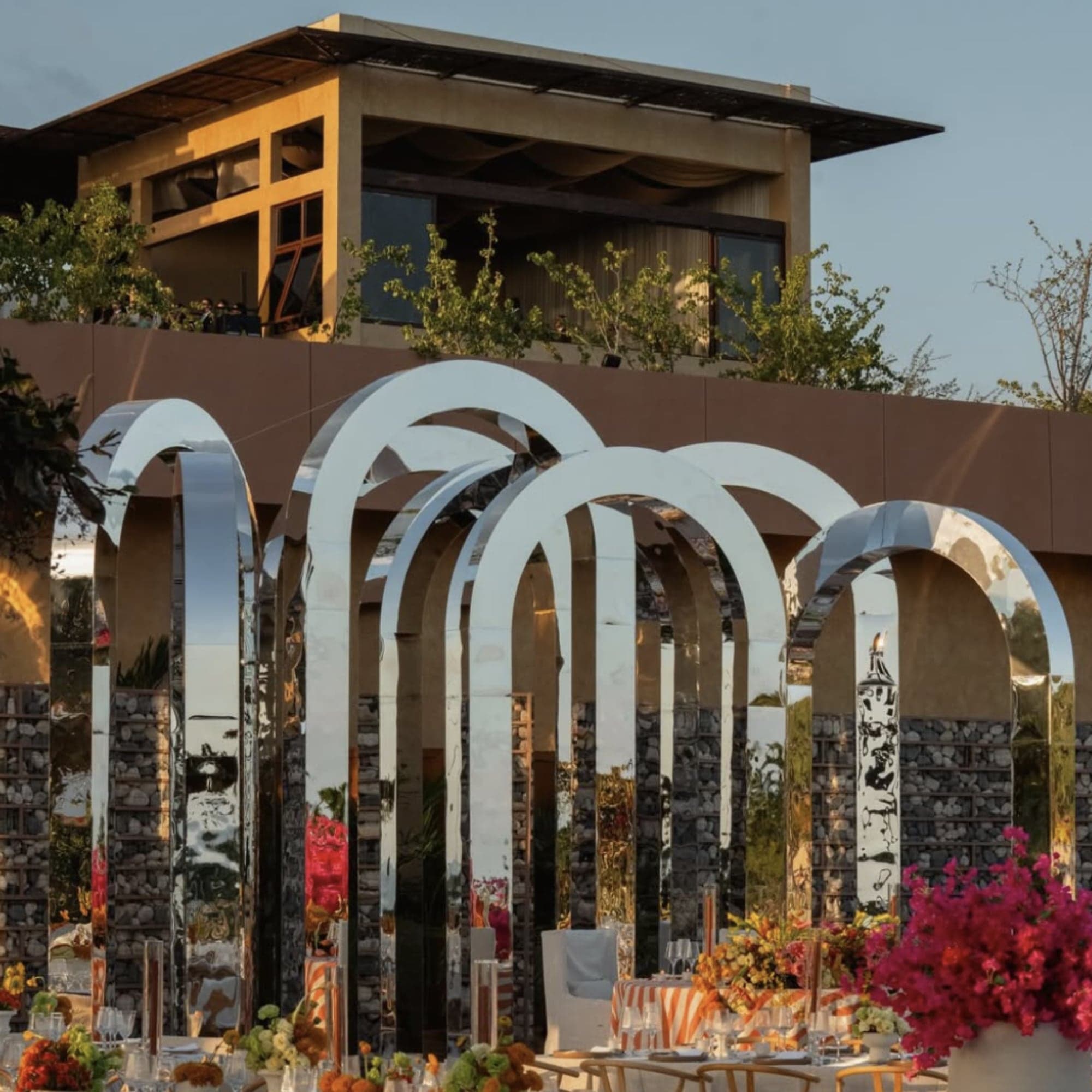 Arcos de acero espejado y flores fucsia junto a una mesa de recepción al atardecer